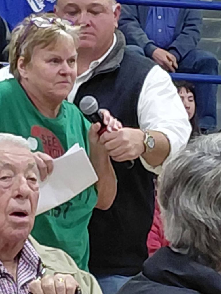 A woman in a green shirt holds a microphone and speaks during a community meeting, expressing concerns about proposed data centers in her area.