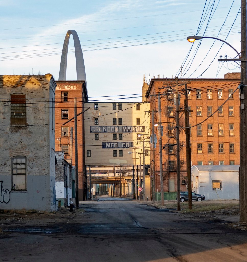 A view of the Crunden-Martin Manufacturing Company complex with the Gateway Arch visible in the background, featuring brick buildings and utility poles lining the street.