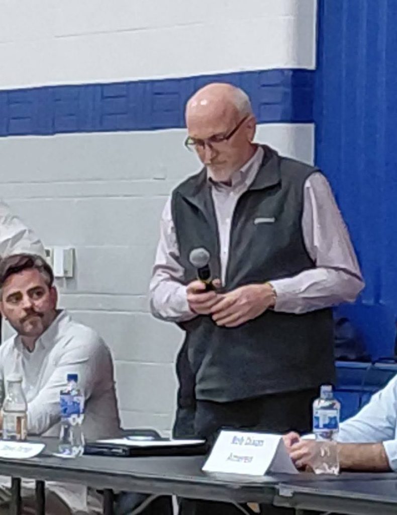 A panel of representatives at a town hall meeting, with one speaker holding a microphone and looking down, while others sit at a table with water bottles and name tags in front of them, against a blue and white backdrop.