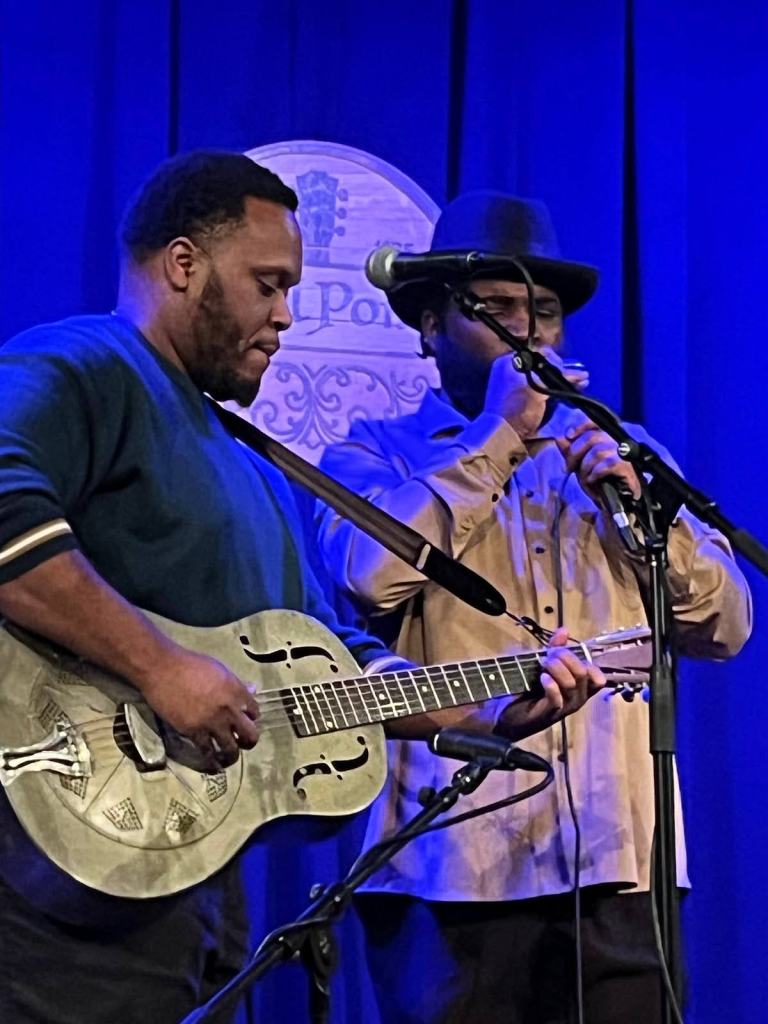 Two musicians perform on stage, one playing a resonator guitar while the other sings and plays harmonica, with a blue backdrop and a guitar-themed sign in the background.
