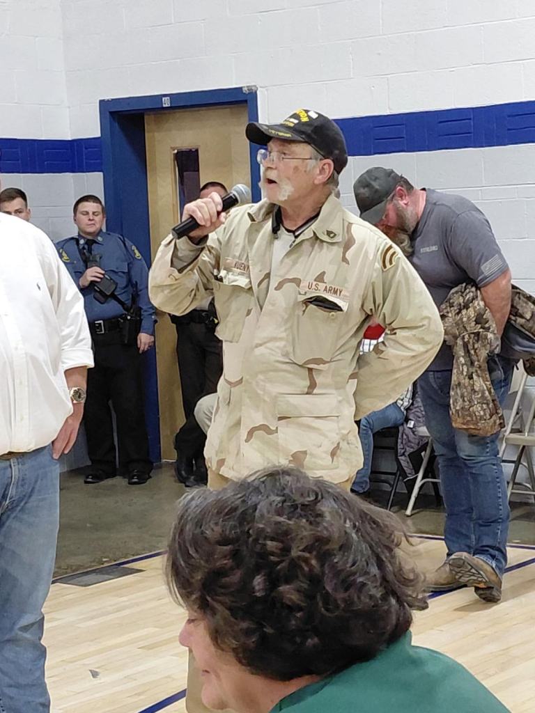 An elderly man in military attire stands at a microphone during a public meeting, addressing attendees. In the background, people are gathered, with some appearing to listen attentively.