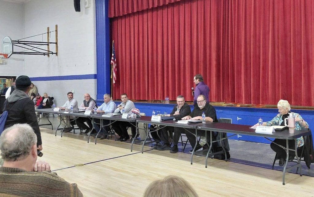 A community meeting at Montgomery City Elementary School featuring a panel of local government representatives seated at a table, discussing concerns about proposed data centers. Attendees are positioned in front of the panel, with red curtains in the background.