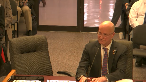 A man in a suit sitting at a table during a meeting or hearing, looking thoughtfully at the proceedings.