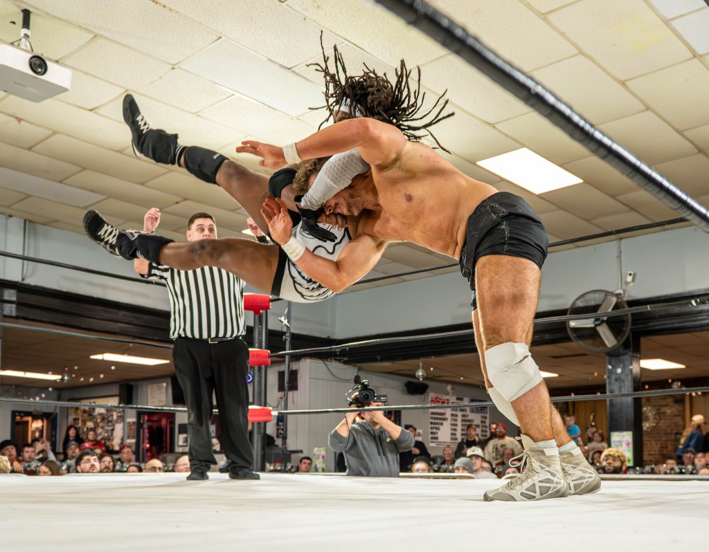 A wrestling match in progress, featuring two athletes engaged in a dramatic move; one wrestler is hoisting the other mid-air in a dynamic pose, while a referee observes the action.