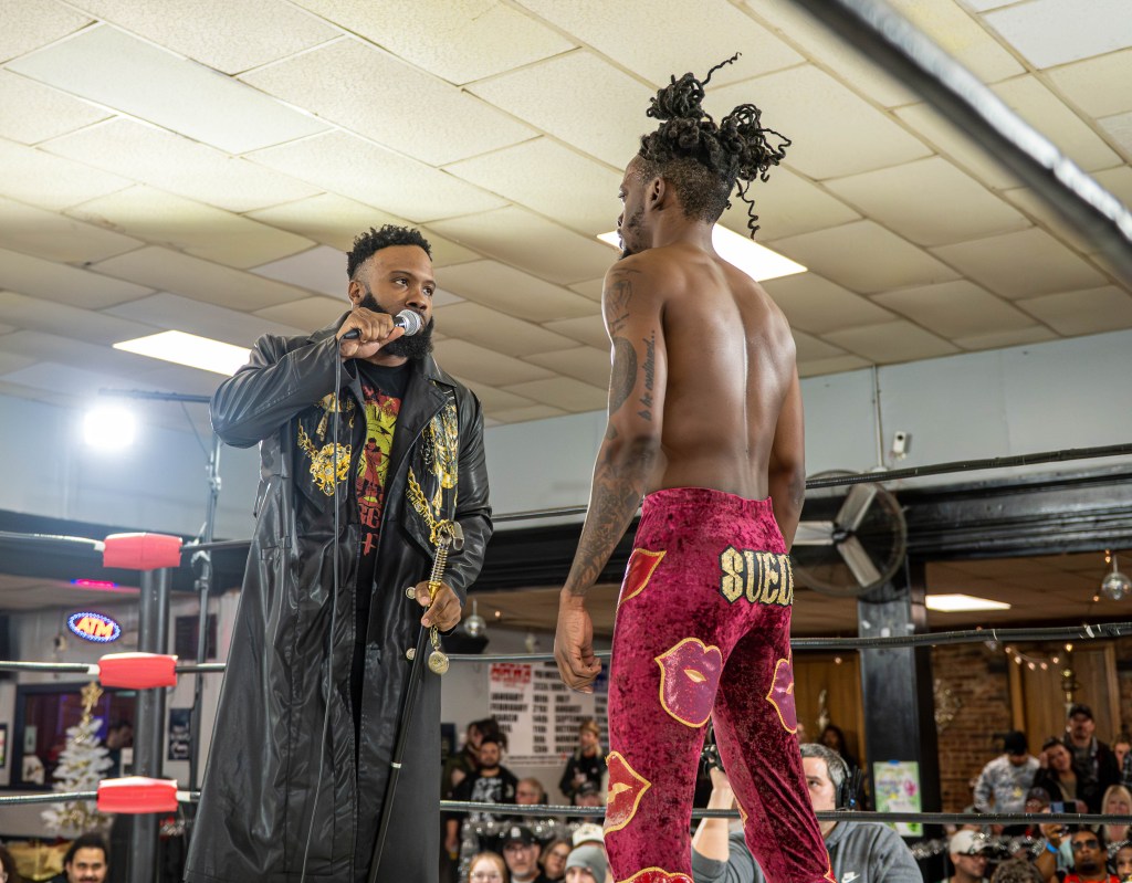 Two professional wrestlers face off in a ring, one speaking into a microphone while the other stands ready. The wrestler speaking wears a black jacket with gold designs, while the other has a bare back and colorful pants with a bold print.