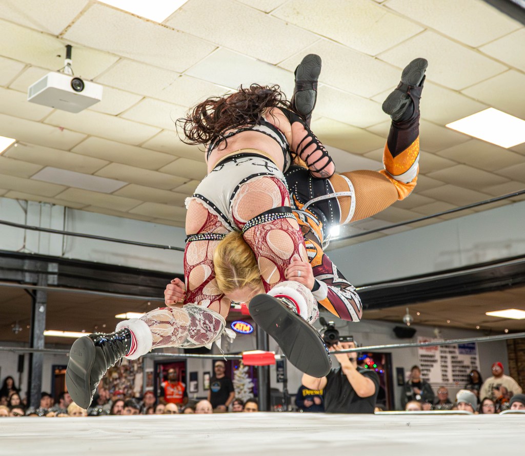Two female wrestlers perform a flying move in a wrestling ring, surrounded by a cheering audience.