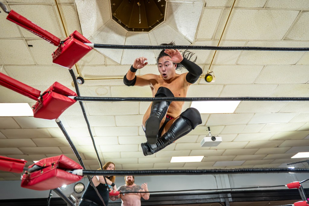 A wrestler leaps from the top rope during a match, showcasing dynamic aerial movement, with spectators visible in the background.
