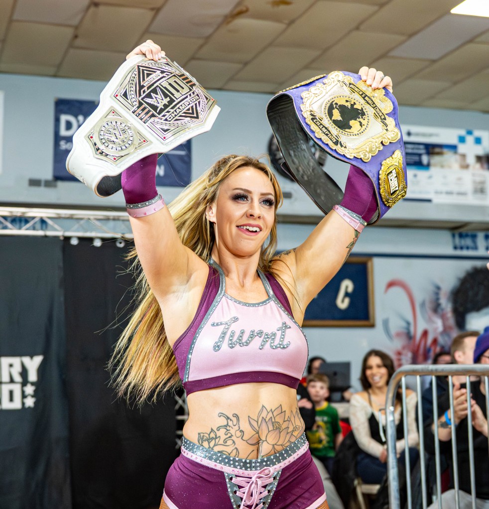 A female wrestler celebrating with two championship belts raised above her head, wearing a purple and black outfit with the word 'Twir' on it, and displaying visible tattoos on her torso.