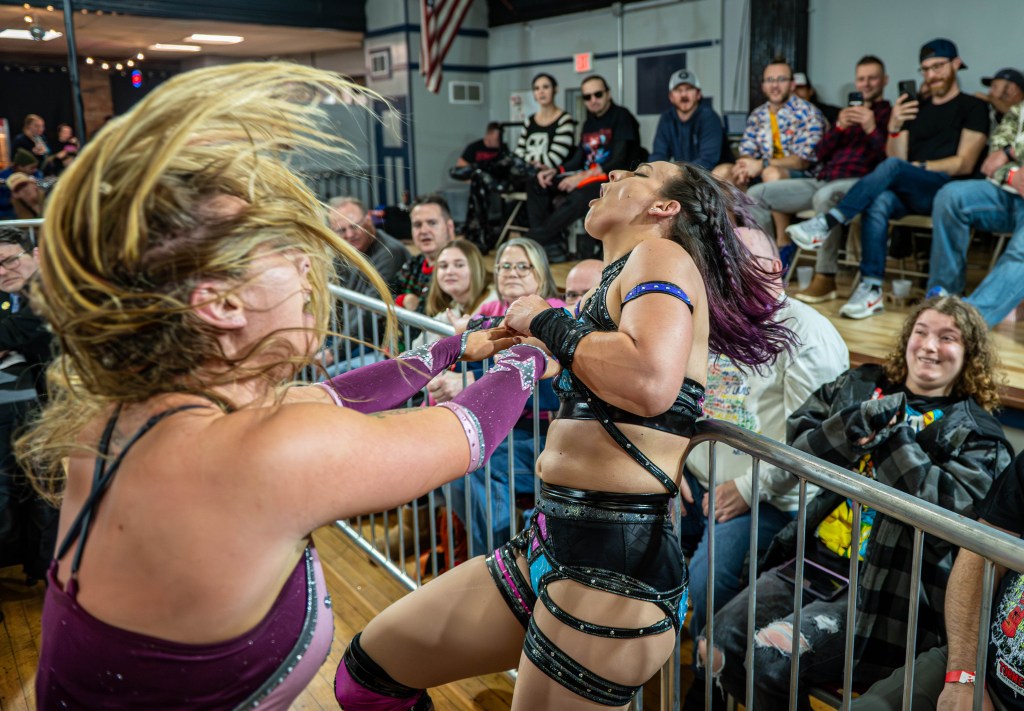 A wrestling match is taking place, featuring two female wrestlers in action. One wrestler is pulling the other towards the barrier while spectators are watching enthusiastically in the background.