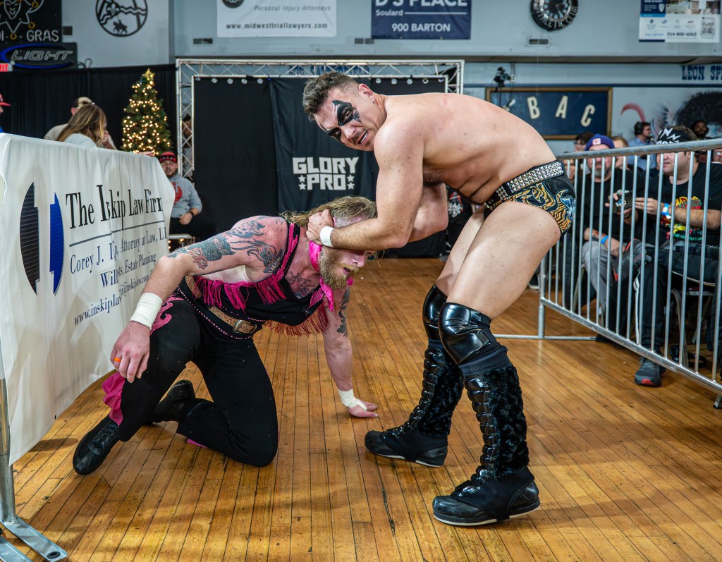 A wrestling match scene with one wrestler in a pink outfit being held in a headlock by another wrestler in black shorts. The audience can be seen in the background, with a festive tree and a banner in the foreground.