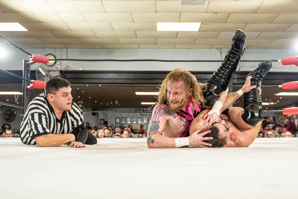 A wrestling match in an indoor ring, showcasing a wrestler with long hair and tattoos applying a hold on an opponent while a referee observes nearby.
