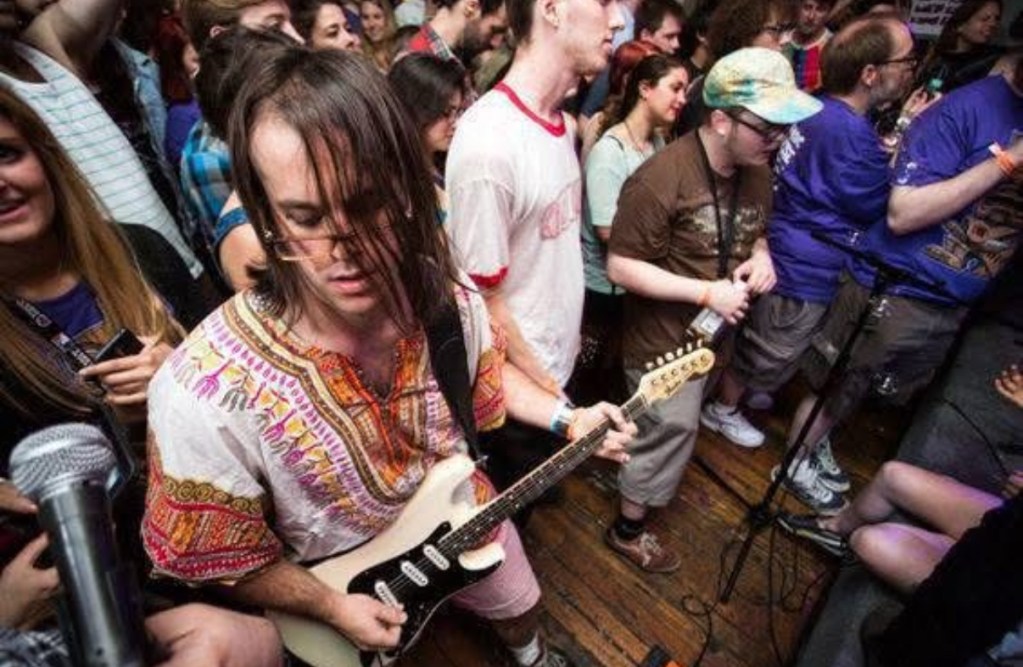 A musician playing electric guitar in a lively crowd at a performance, wearing a colorful embroidered shirt, surrounded by fans enjoying the show.