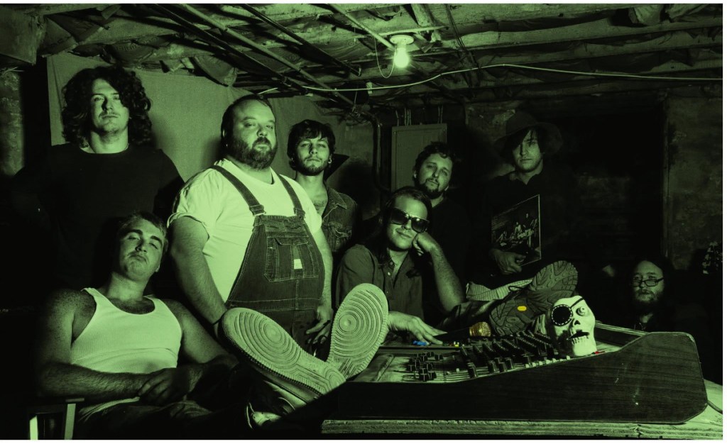 Group of men posing in a dimly lit basement with musical equipment and vintage decor.