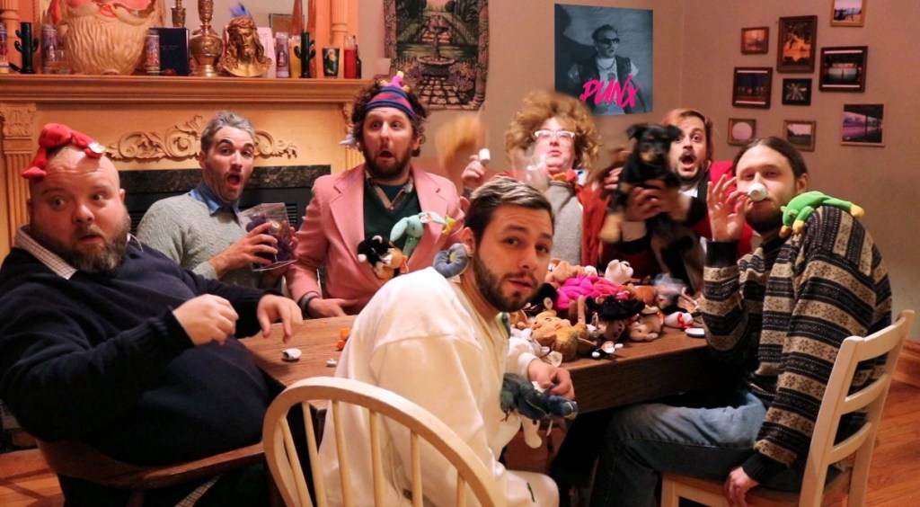 A group of eight people gathered around a table, making funny faces and holding various items, including stuffed toys and drinks, in a playful setting.