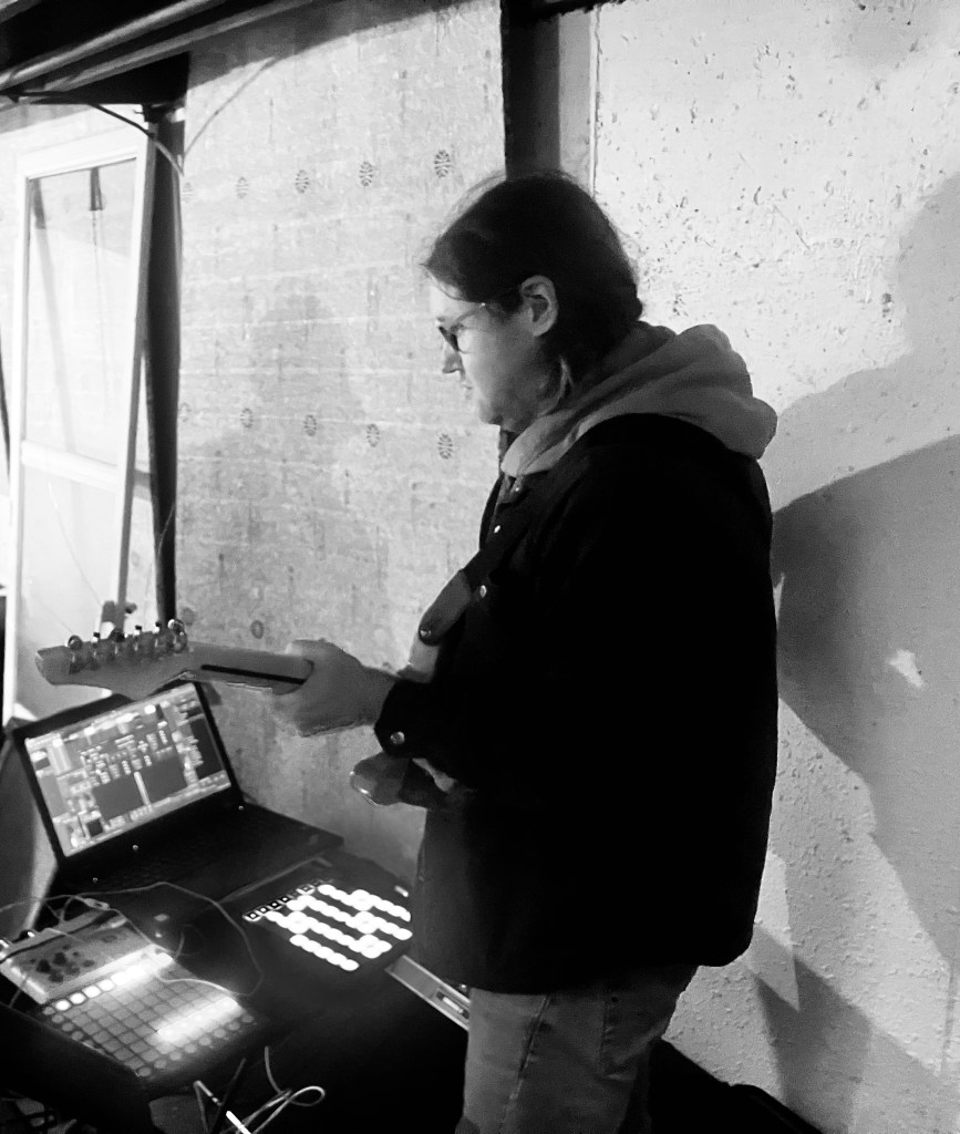 A musician holding a guitar, focused on a laptop with music software and a MIDI controller, set against a textured wall.