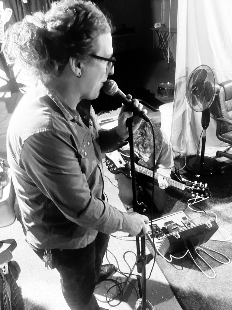 A musician singing into a microphone while standing on stage, with another musician playing guitar on the ground in the background.