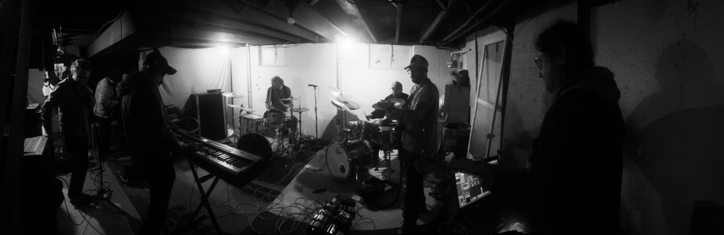 A black and white image of a band rehearsing in a basement with various instruments, including a keyboard, drums, and guitars. Musicians are positioned around the room engaged in playing music.