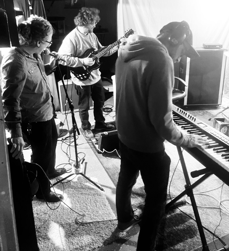 A black and white image of three musicians performing in a studio setting. One musician is singing into a microphone, another is playing an electric guitar, and the third is playing a keyboard.