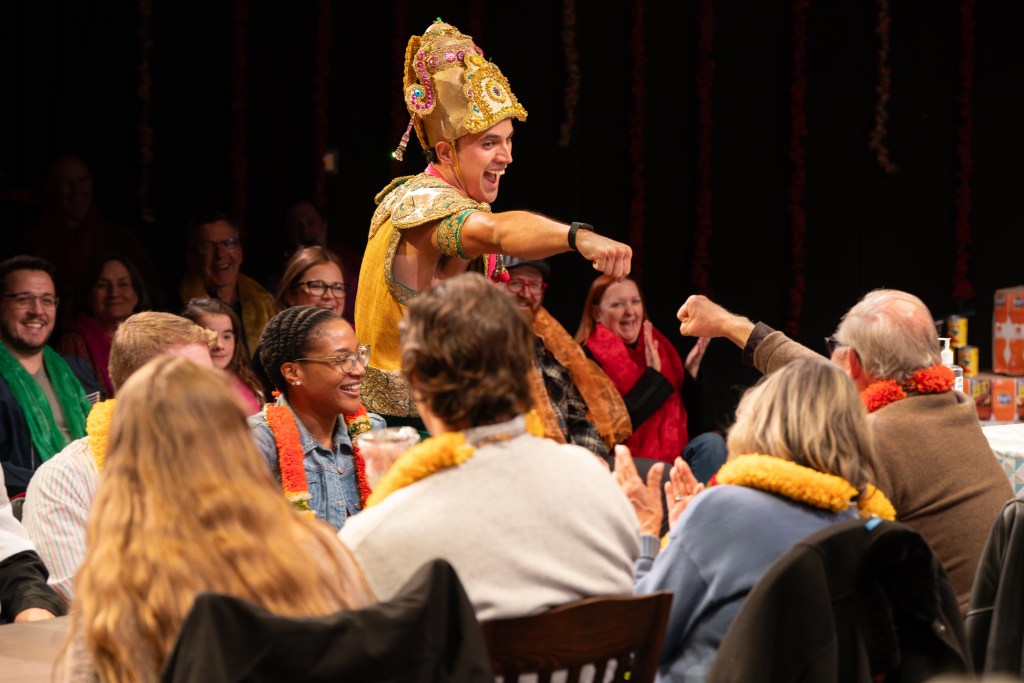 A performer dressed in colorful traditional attire interacts enthusiastically with an audience during a lively event. The audience is engaged and smiling, with some applauding and others wearing festive garlands.