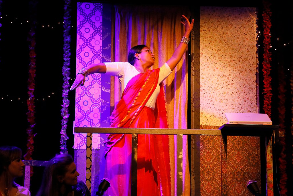 A performer in a red sari striking a dramatic pose on stage, with a colorful backdrop and audience members visible in the foreground.