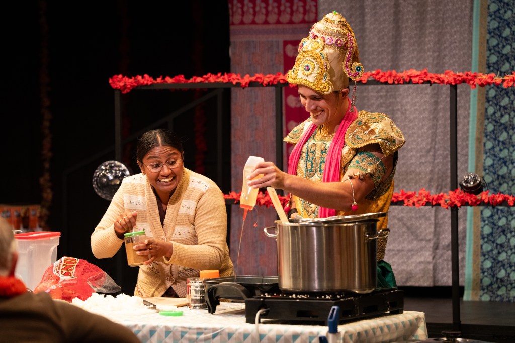 A woman smiles while holding a jar, engaged with a man dressed in traditional Indian attire, who is stirring a pot on a stove during a cooking demonstration.