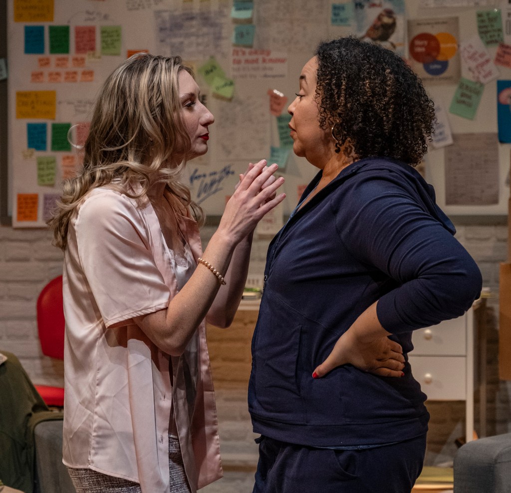Two women engaging in an intense conversation in a room filled with notes and papers on the walls.
