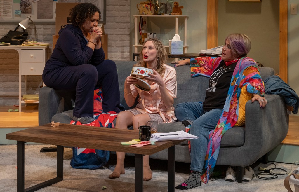 Three women sitting on a couch in a living room setting, engaged in conversation, with one woman holding a bowl. The room has a cozy but cluttered atmosphere.