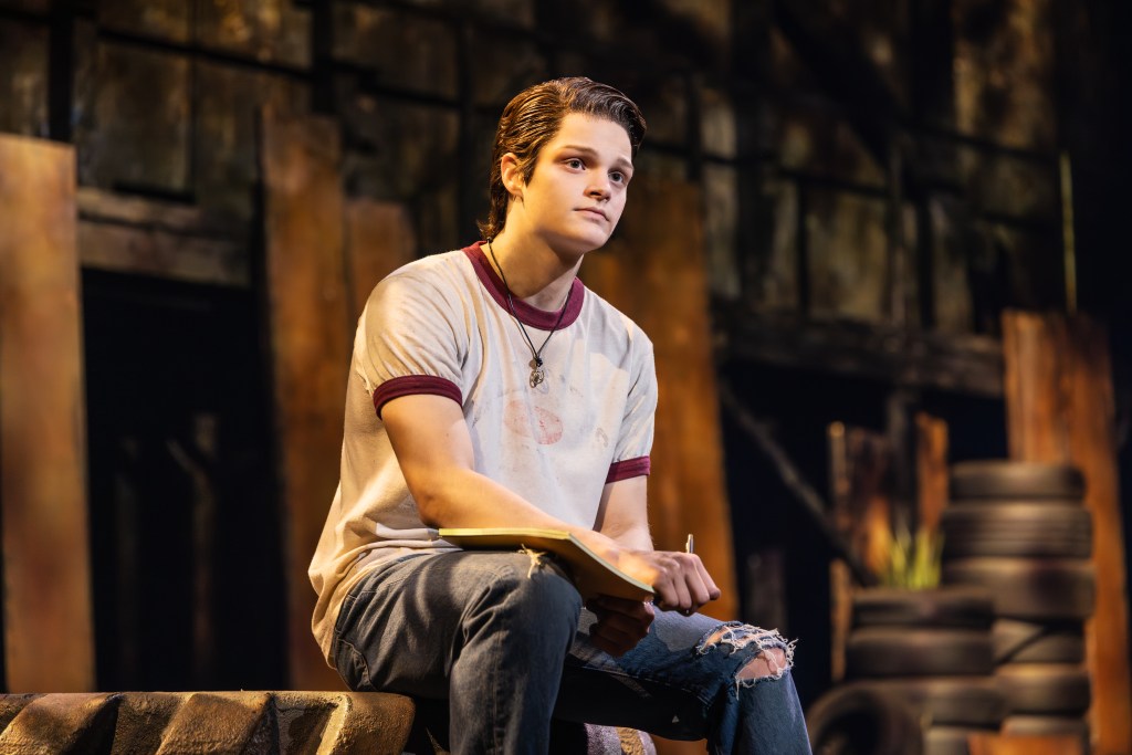 A young actor portraying Ponyboy Curtis, sitting on a tire in a theatrical setting, holding a notebook and looking contemplative.
