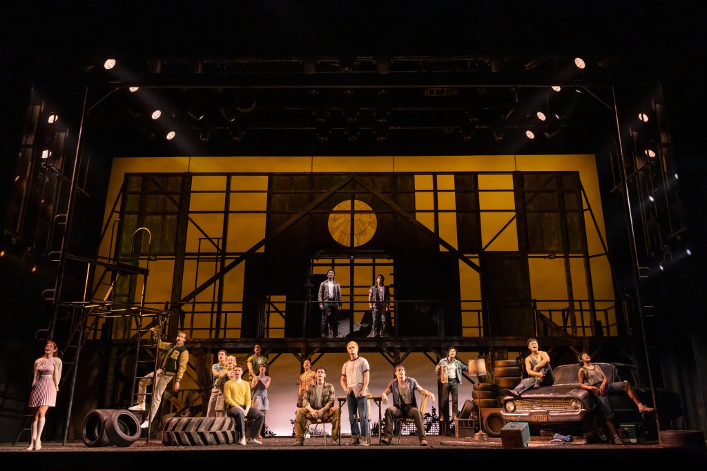 A scene from the musical 'The Outsiders' featuring a cast standing and sitting on stage, with a stylized set design that includes industrial elements and a warm-colored backdrop.