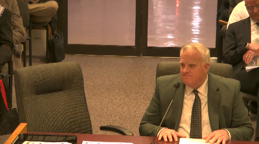 A man in a suit sitting at a table during a meeting or hearing, with another person visible in the background and a window reflecting light.