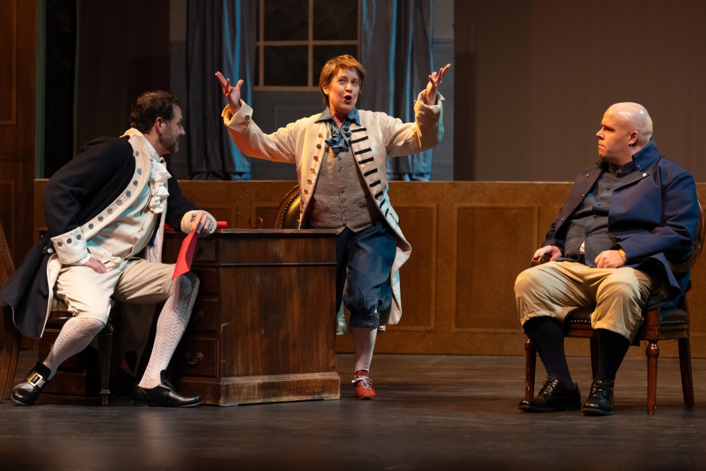 Three actors performing on stage, with one energetically gesturing while leaning toward the other two seated individuals, who are observing intently.