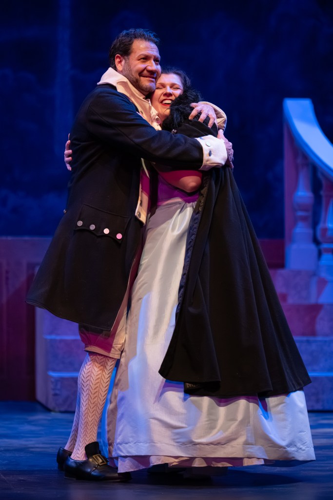 Two actors embracing on stage during a performance, dressed in period costumes with a colorful backdrop.