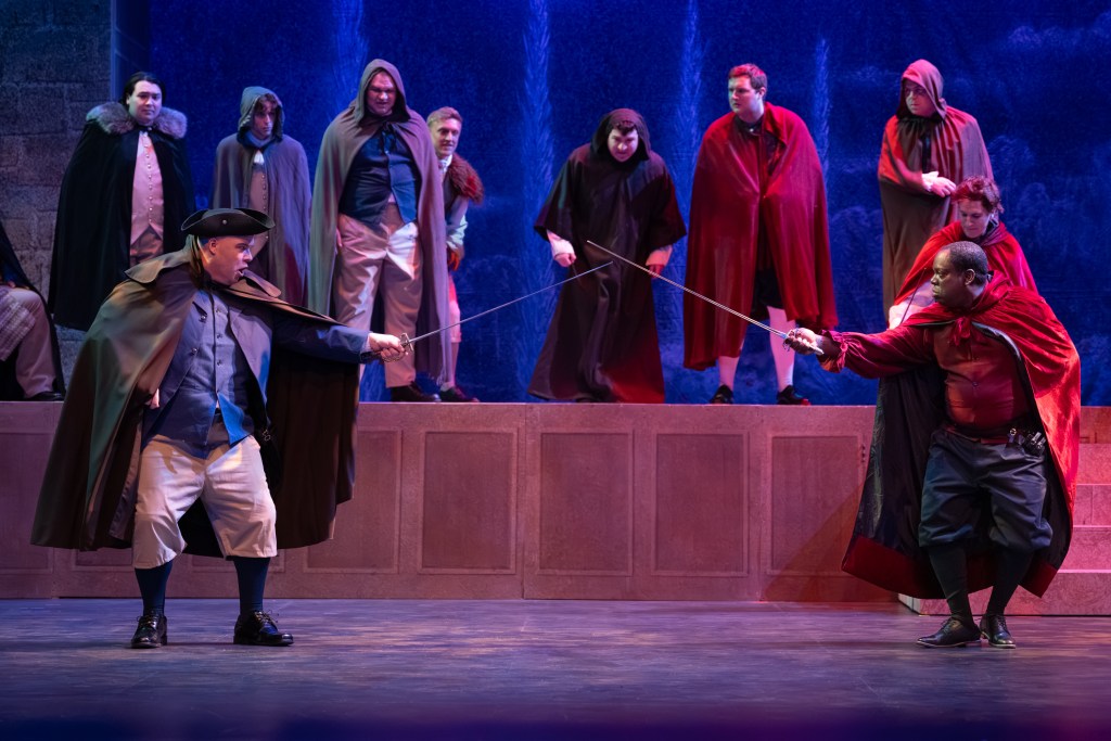 A staged sword duel between two actors, one in a tricorn hat and cape, and the other wearing a red cape, with several cloaked figures watching from the background.