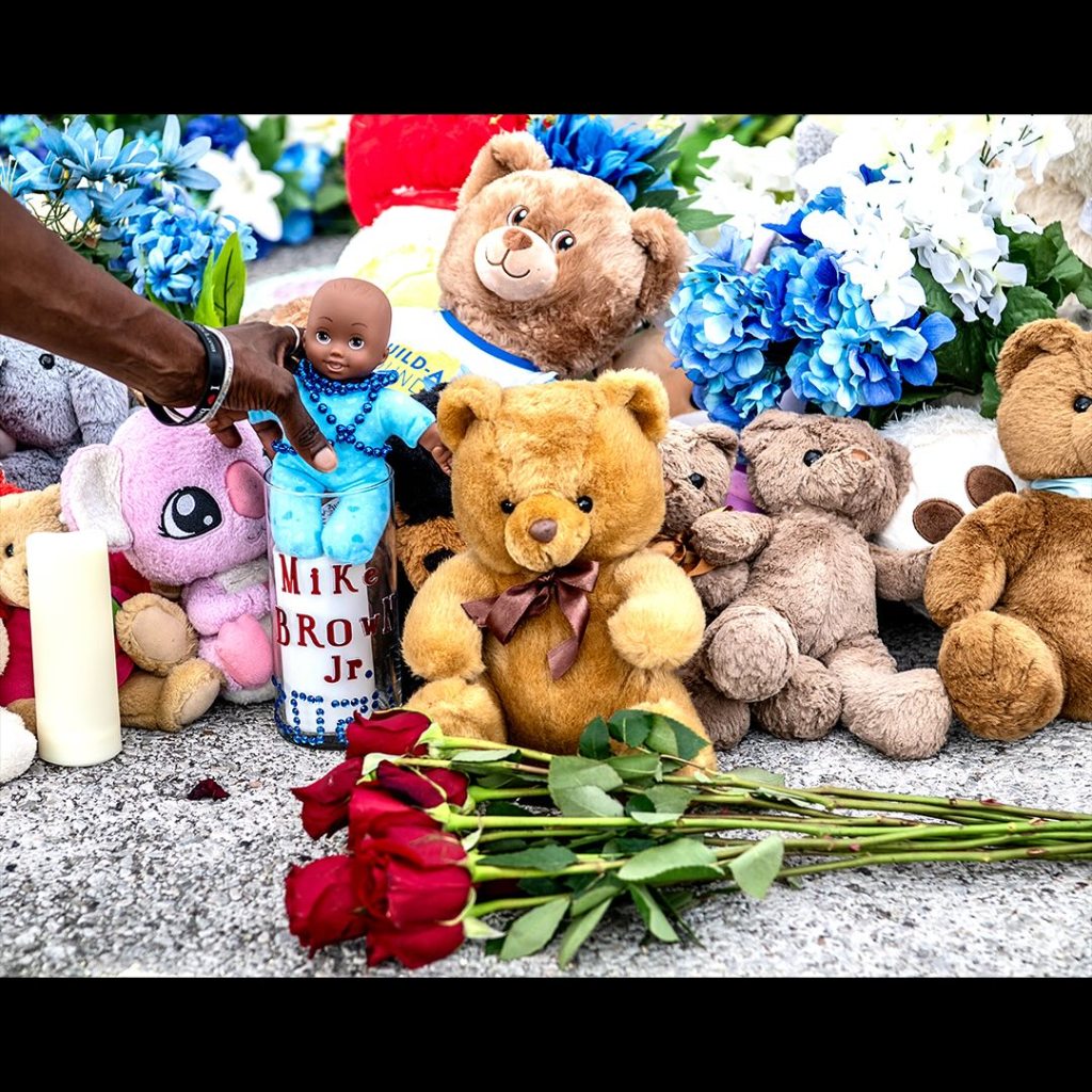 A memorial featuring various teddy bears, a baby doll, a candle, and red roses, with a hand placing a doll beside a candle that has 'Mike Brown Jr.' written on it.