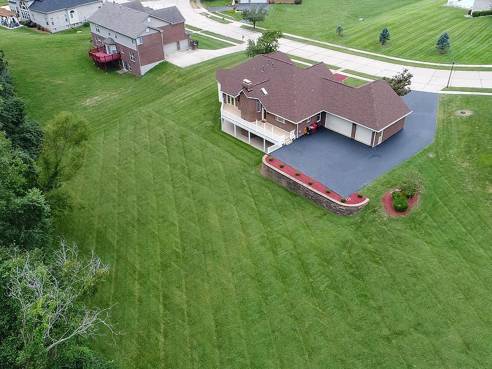 Aerial view of a spacious residential property featuring a large house with a brown roof and a paved driveway, surrounded by well-maintained green lawns and landscaped flower beds.