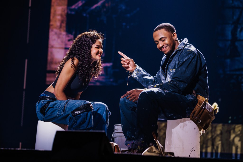 A male and female performer sitting on stage, sharing a playful moment while smiling and engaging with each other.