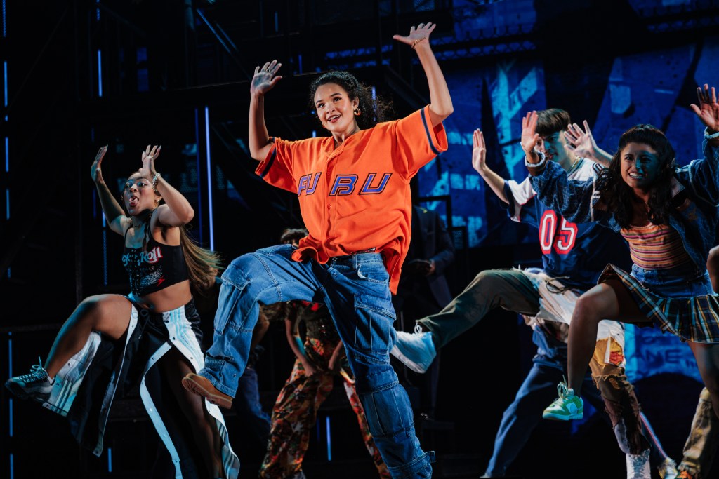 A group of young dancers performing energetically on stage, showcasing various dance moves. The central dancer is wearing an orange oversized shirt and denim pants, while others sport a mix of casual outfits. The backdrop features vibrant lighting and a modern urban aesthetic.