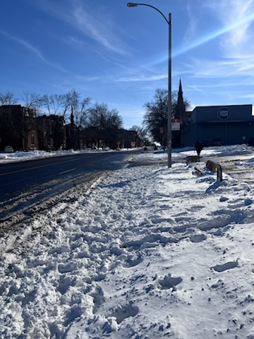 A snowy street scene on a clear day, with snow piled along the edge of the road and a few trees in the background. A church spire is visible in the distance.