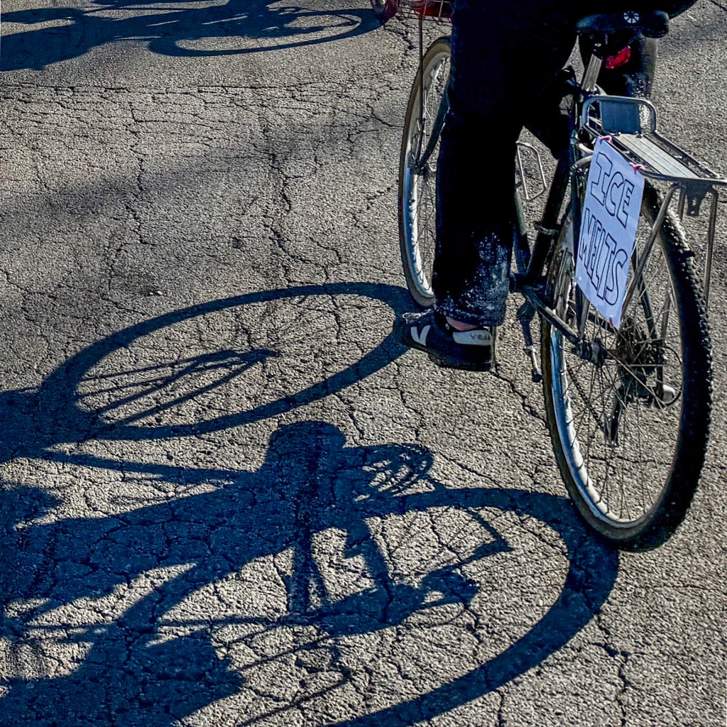 Close-up of a bicycle tire casting a shadow on cracked pavement, with a sign reading 'ICE MILLS' attached to the bike.
