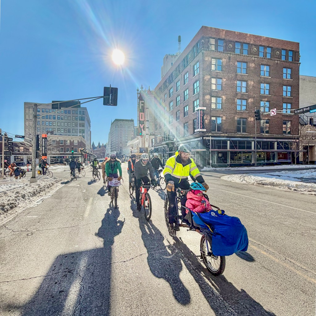 A group of cyclists riding through a snowy city street on a sunny day. Notable buildings and traffic signs can be seen in the background.