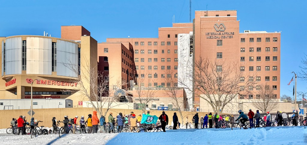 A group of people on bicycles gathered in the snow in front of the Veterans Affairs Medical Center, with the emergency entrance visible on the left side of the image.