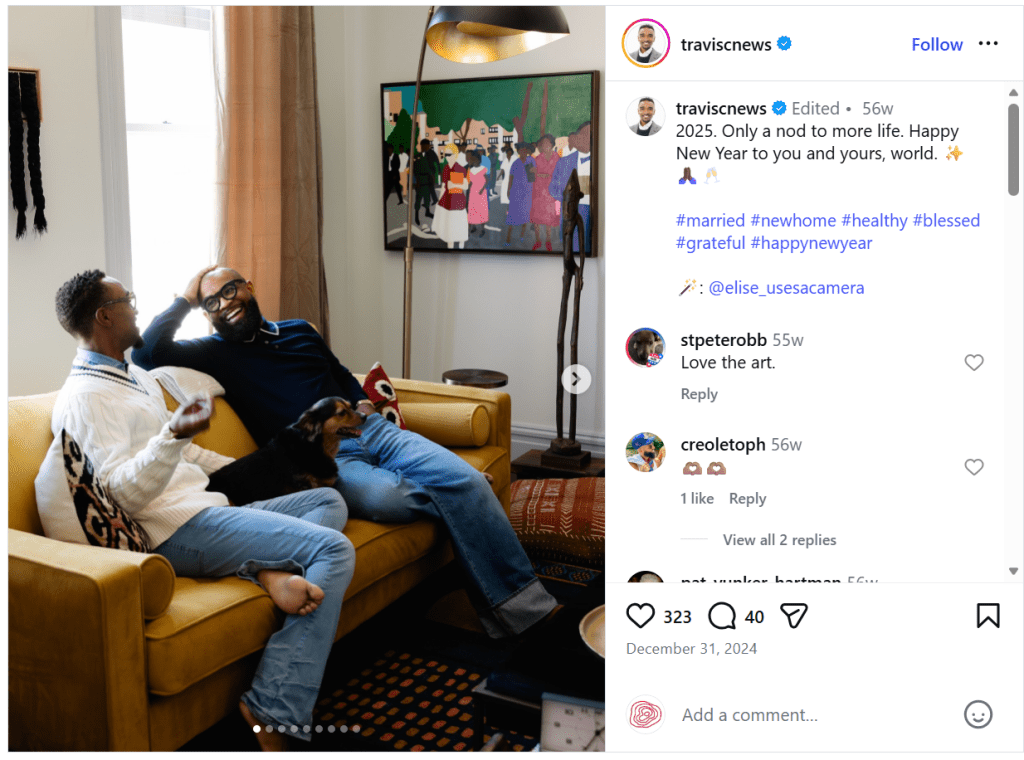 Two men relax on a yellow couch in a cozy living room, engaged in conversation with a small dog lying beside them. A colorful piece of artwork hangs on the wall in the background.