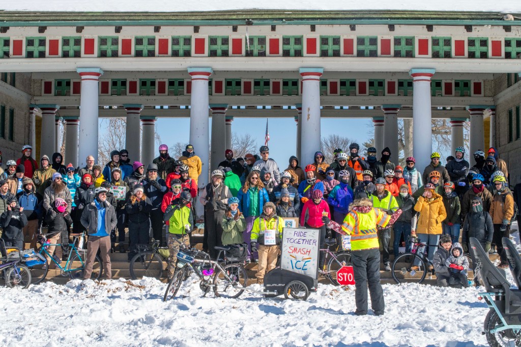 A large group of people, many wearing winter clothing and helmets, gather on steps covered in snow in front of a historic building. Several bicycles are parked in front, and a person in bright yellow attire holds a sign that promotes unity and biking activities.