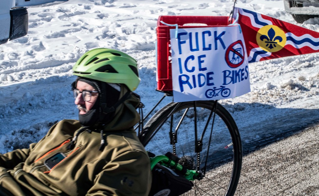 A cyclist wearing a green helmet and glasses, sitting on a bicycle on a snowy road, with a sign attached to the bike that says 'FUCK ICE RIDE BIKES'.