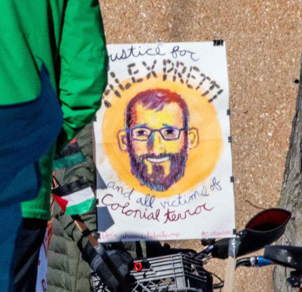 A poster featuring a colorful portrait of a bearded man with glasses, surrounded by the text 'Justice for Alex Preti and all victims of Colonial Terror.' The poster is held in a crowd, with flags and a bicycle visible in the foreground.