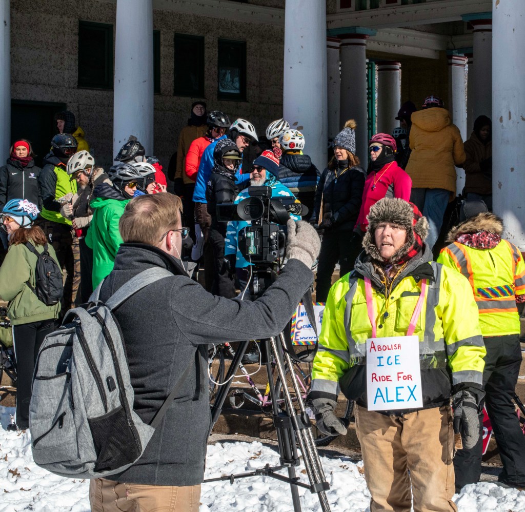 A group of people gather outdoors in winter attire, some wearing helmets and standing near bicycles, as a person in a bright yellow jacket holds a sign that reads 'Abolish ICE Ride for Alex.' A man is filming the group with a camera.