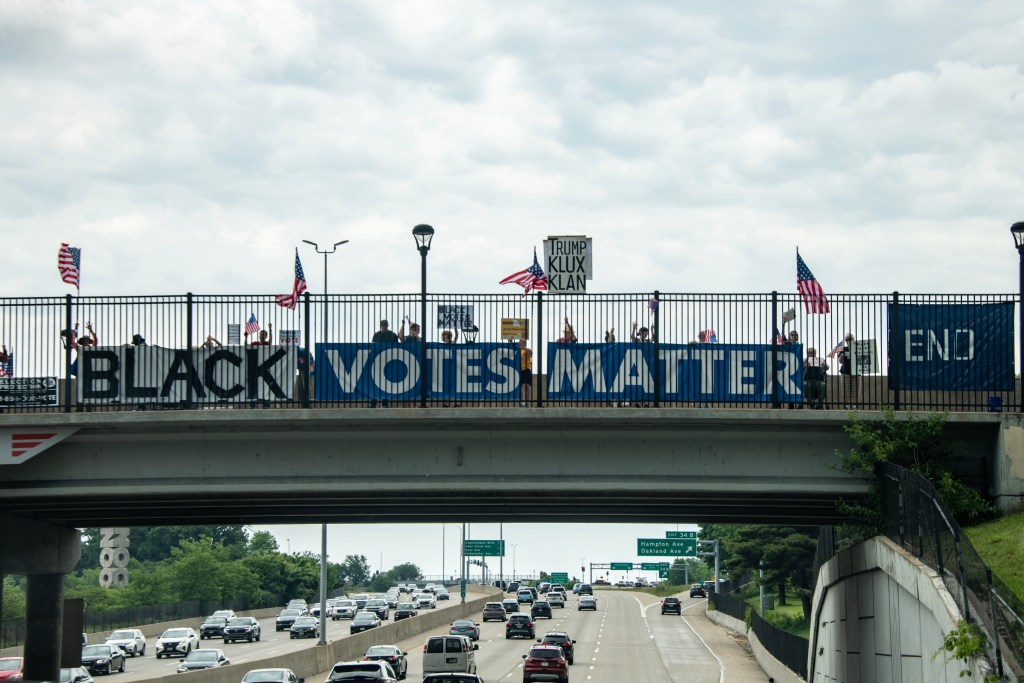 A large banner reading 'BLACK VOTES MATTER' displayed on a bridge with people holding flags and signs, while cars pass underneath.