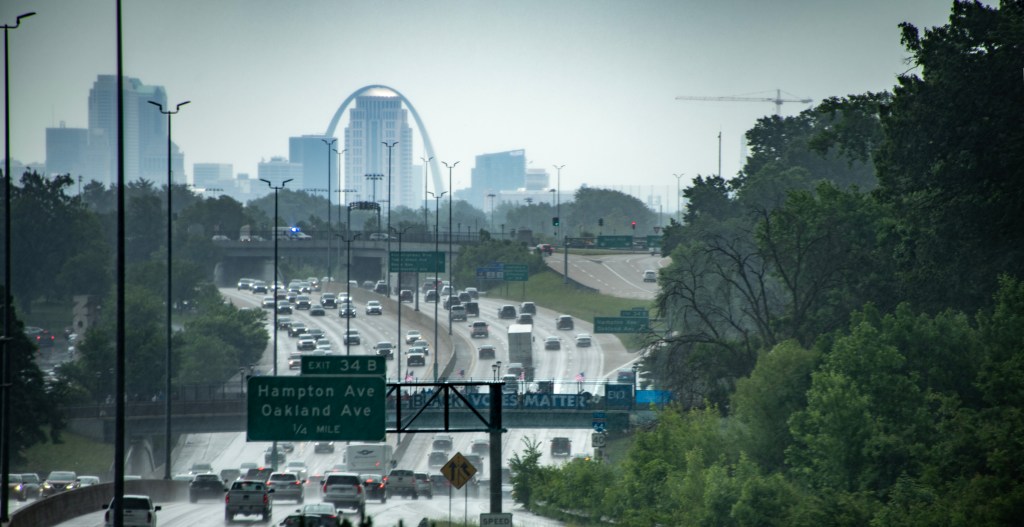 A busy highway scene with cars driving under cloudy skies, leading towards a city skyline featuring high-rise buildings and an arc structure in the background.