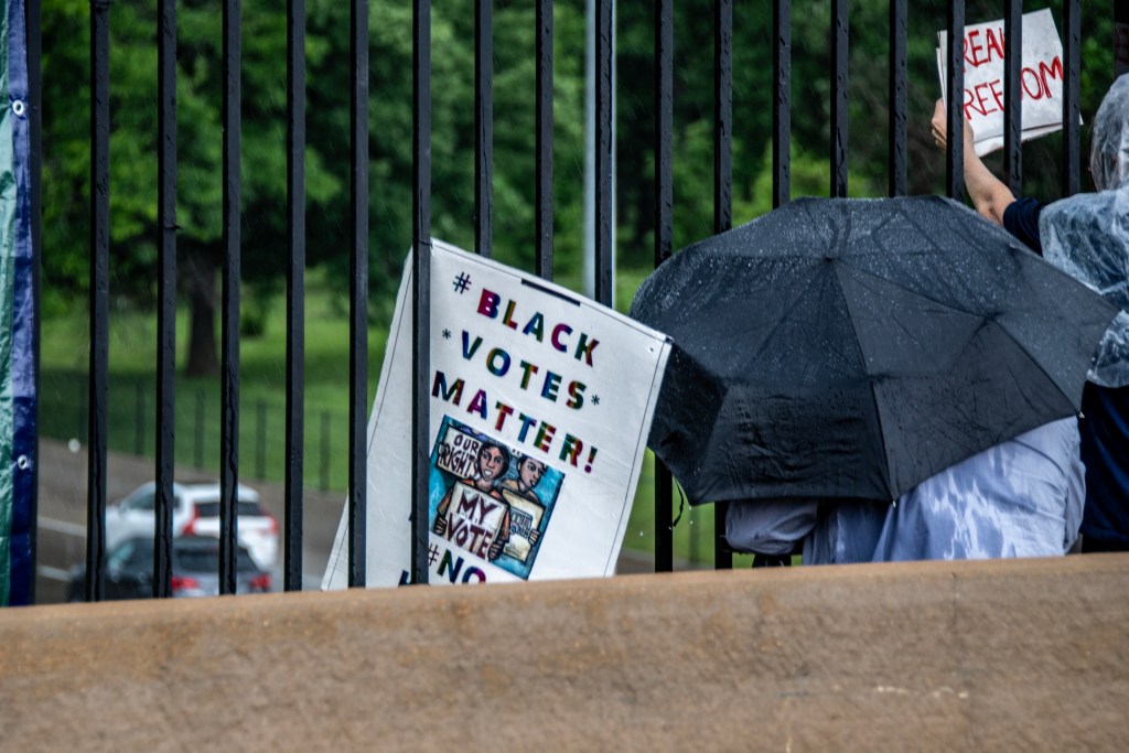 Protesters holding signs advocating for voting rights and equality, under black umbrellas in the rain.