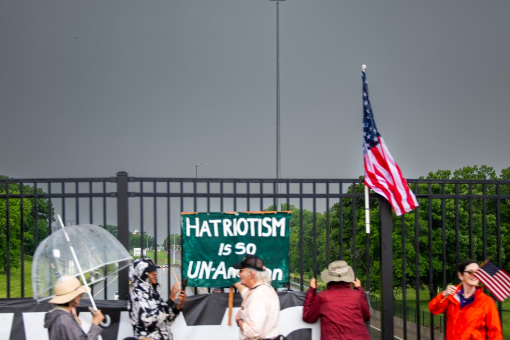 Protesters holding a sign that reads 'Hatriotism is so UN-American' with an American flag in the background, set against a stormy sky.